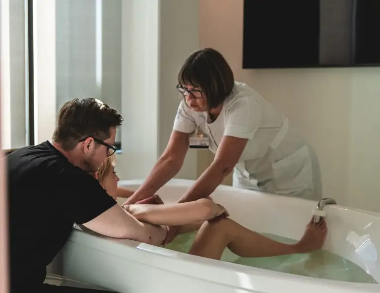 Kvinde der føder i badekar på fødestue med parner og jordemoder på hver sin side